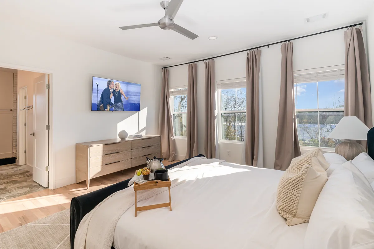 A sunlit bedroom featuring a plush bed adorned with decorative pillows, a TV displaying a cheerful image, and expansive windows that fill the space with natural light. The room exudes a peaceful and cozy atmosphere, perfect for relaxation and unwinding.