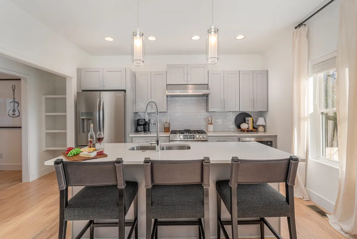 A bright, inviting kitchen with an island counter and bar stools is bathed in natural light from a large window. Modern kitchen appliances and uncluttered surfaces create a welcoming setting suitable for cooking, entertaining, or casual family meals.