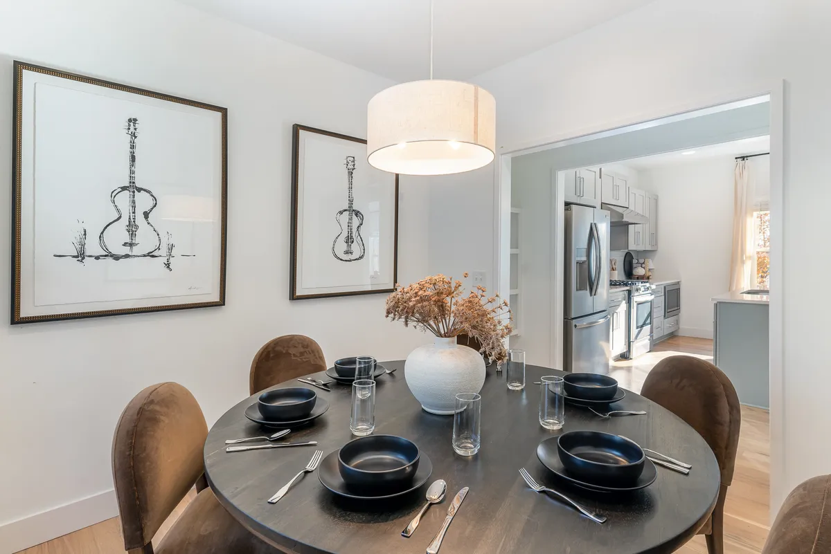 A warm dining area with a spacious table set for gatherings, flanked by two framed guitar illustrations and a vase with dried flowers. The kitchen is visible through an open entrance, suggesting easy movement between dining and culinary spaces.