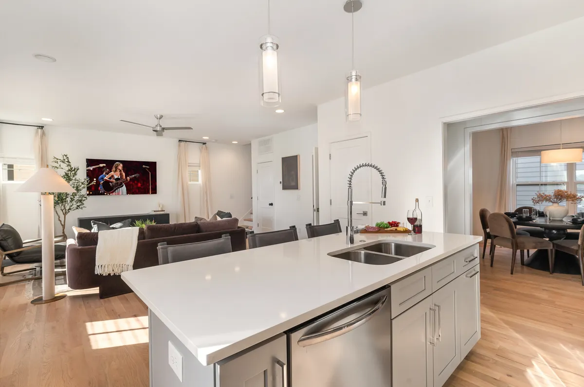 This image features an open-concept living space with a seamless flow between the kitchen and living room. The island kitchen includes a sink and modern cabinetry, while the living room offers comfortable seating and a wall-mounted smart TV. Large windows let in abundant natural light, emphasizing the airy, inviting layout.
